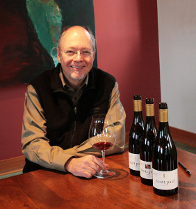 A photo of a smiling man in glasses next to bottles of wine.