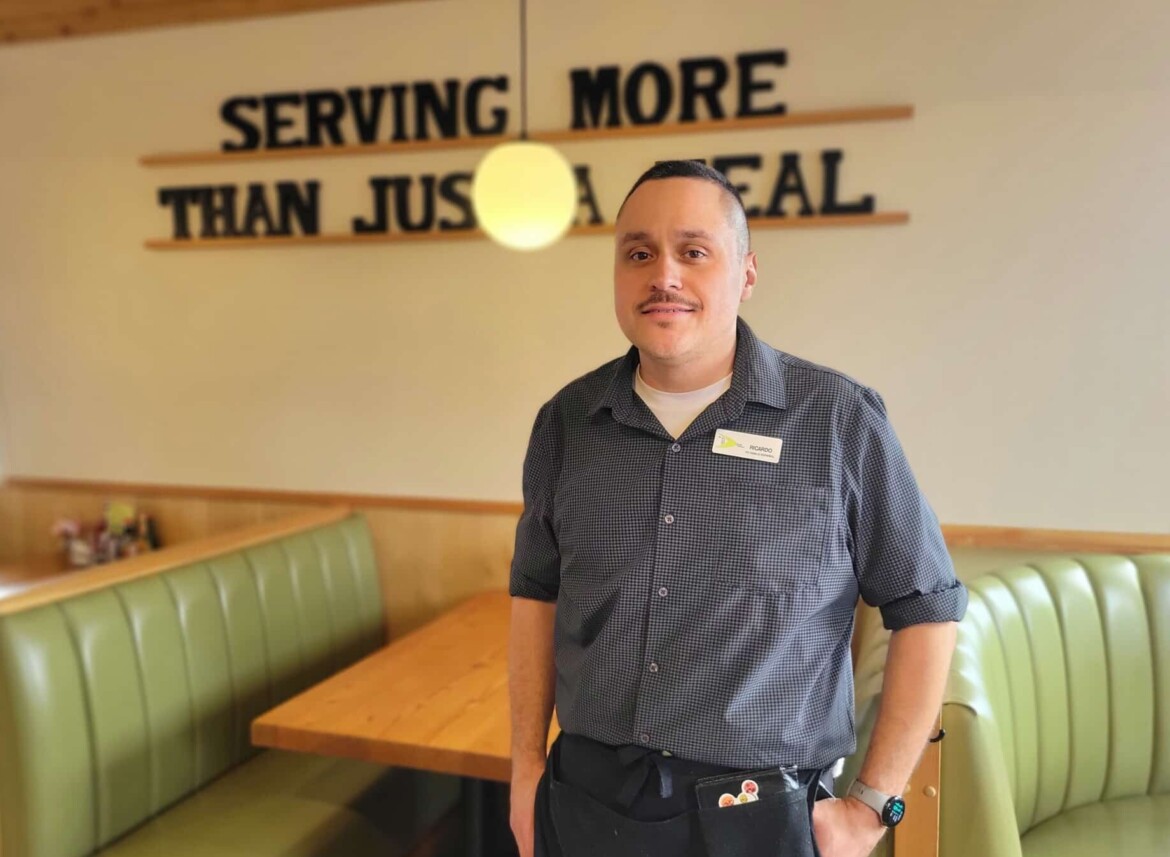 A photo of a man with his hands in his pockets in front of a table at a diner.